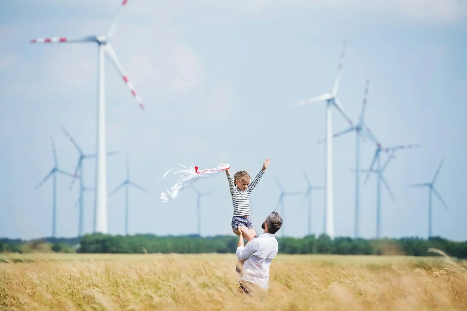 En vuxen lyfter ett barn i en sommaräng med vindkraftverk i bakgrunden – symbol för framtid, glädje och hållbar energi.
